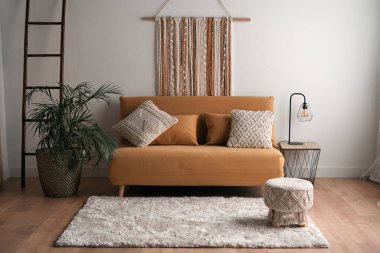 Pastel renklerde oturma odasının içinde rahat bir kanepe, halının yanına konmuş yastıklar, saksılar ve dairedeki lambalar.