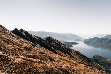 Dar, yılan şekilli toprak yol dağın tepesine, büyük mavi gölün yakınındaki çalıların ve bitkilerin arasına doğru gidiyor. Kayalık dağlar ve mavi gökyüzü arasında sakin bir su var.