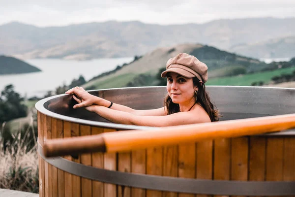 blue-eyed caucasian young girl with brown hat and bracelet on her wrist and crossed hands inside the hot water of the round jacuzzi in the middle of the vegetation and mountains, te wepu pods akaroa