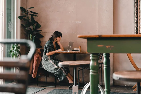 young caucasian woman with disheveled hair in long dress sitting on metal chair by wall typing laptop at wooden table in quiet coffee shop, the mylk bar, new zealand - Lifestyle concept