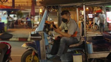 Tuk tuk sürücüsü sürüşler arasında dinlenirken akıllı telefon kullanıyor. Dışarıda maskeli bir adam var. - 4K Yatay Görüntü