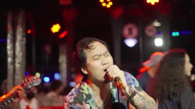 Dövmeli Asyalı grup lideri, arka planda komik pozlar veren kadın grup üyesiyle şarkı söylüyor. Bangkok 'ta Gece Sokak Konseri. Kapat - 4K Yatay Görüntü