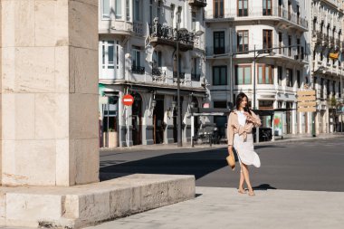 full length of brunette woman in skirt walking along urban street clipart