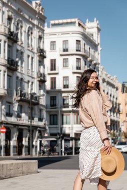 brunette woman with straw hat looking at camera on street clipart