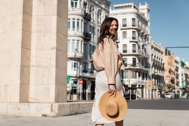 cheerful woman with straw hat walking on windy street and looking at camera clipart