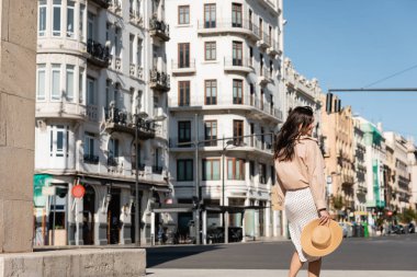 back view of woman in skirt holding straw hat while walking on street