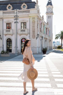 full length of stylish woman looking at camera near crosswalk and building with clock tower