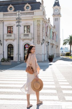 full length of stylish woman looking at camera near crosswalk and building with clock tower clipart