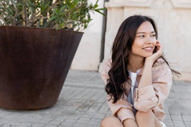 cheerful woman sitting on street near huge flowerpot and looking away 