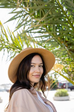 portrait of brunette woman in straw hat smiling at camera near blurred palm tree