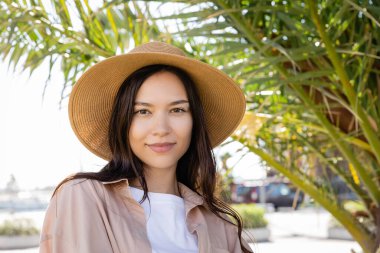 portrait of positive brunette woman in straw hat looking at camera near blurred palm leaves