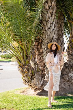 full length of happy woman in straw hat and skirt standing near giant palm tree