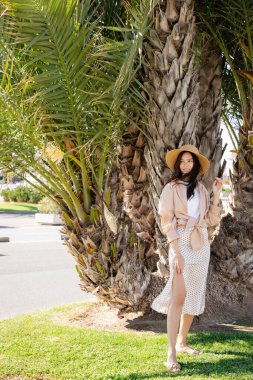 full length of happy woman in straw hat and skirt standing near giant palm tree clipart