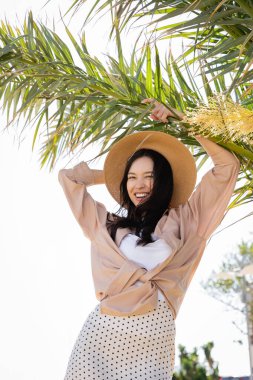 stylish and cheerful woman in straw hat laughing under palm tree