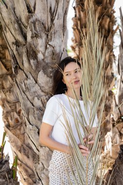 happy brunette woman in white t-shirt standing near palm tree trunks 