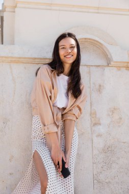 Akıllı telefonlu neşeli kadın beyaz duvarın yanında duruyor ve kameraya gülümsüyor.