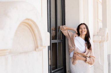 Bej gömlekli neşeli esmer kadın binanın yanında eli başının arkasında duruyor.