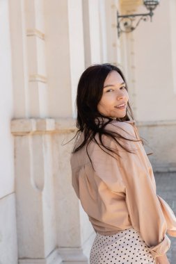 Bej tişörtlü neşeli esmer kadın şehir sokağında kameraya bakıyor.