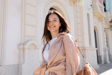 pleased brunette woman in beige shirt smiling at camera near blurred building