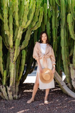 full length of happy and stylish woman looking at camera near giant succulents clipart