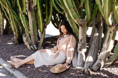full length view of brunette woman in skirt sitting under giant cacti near straw hat