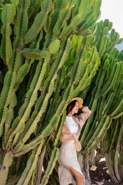 brunette woman in straw hat smiling near giant cacti in park