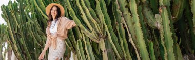 cheerful woman in trendy clothes and straw hat near huge cacti, banner