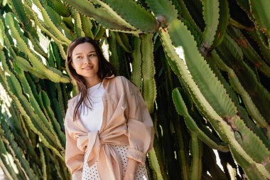 low angle view of brunette woman in shirt smiling near giant cacti