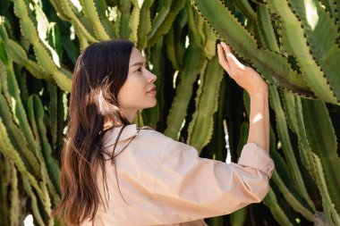 side view of pretty woman in beige shirt touching green succulents in park