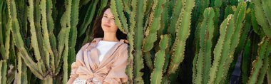 brunette woman in beige shirt resting near green succulents in park, banner