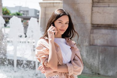 Bej gömlekli kadın cep telefonuyla konuşuyor fıskiyenin yanında bulanık arka planda.
