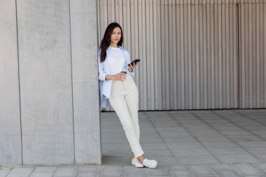 Gri duvarın yanına gitmek için elinde akıllı telefon ve kahveyle bekleyen esmer kadın.
