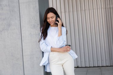 Elinde kağıt bardakla gülümseyen esmer kadın gri duvarın yanında cep telefonuyla konuşuyor.