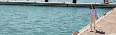 full length of brunette woman with paper cup walking along pier, banner
