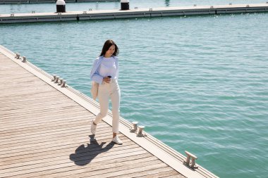 full length of woman in white pants walking with takeaway drink along pier