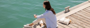brunette woman looking at water while sitting on pier with takeaway drink, banner