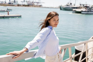 smiling woman in blue shirt looking at camera while standing on pier in seaport