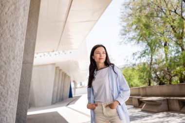 Esmer kadın elleri ceplerinde bina yakınında duruyor ve başka tarafa bakıyor.