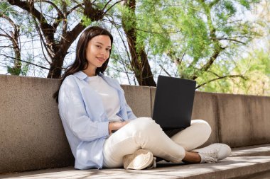 Şehir sokağında dizüstü bilgisayarla oturan mutlu kadın kameraya bakıyor.