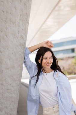 happy woman in blue shirt standing with hand above head near grey wall