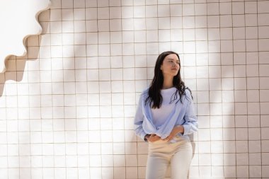 brunette woman in blue shirt standing near white tiled wall and looking away