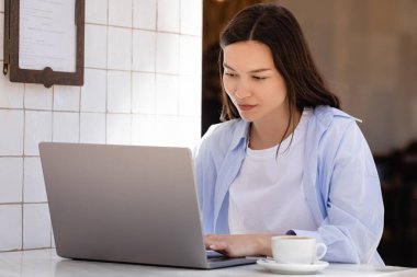 woman in blue shirt typing on laptop near coffee cup on cafe terrace