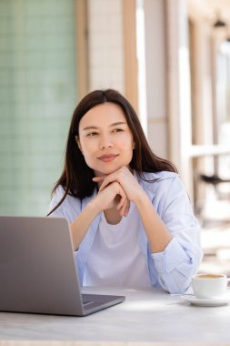 smiling and dreamy woman looking away near laptop on cafe terrace