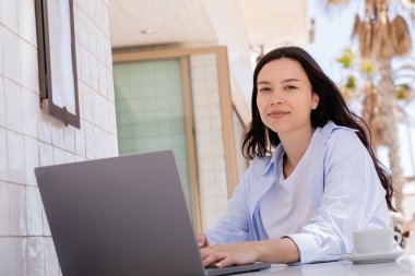 brunette woman smiling at camera while typing on laptop on summer terrace