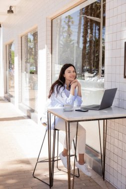 happy woman looking at camera while sitting on cafe terrace near laptop and smartphone