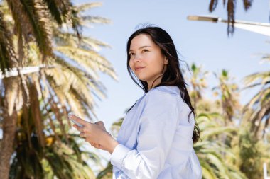 Mavi gömlekli gülümseyen kadın parkta akıllı telefonu tutarken gözlerini kaçırıyor.