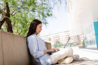 side view of smiling woman sitting with crossed legs on city street and typing on laptop