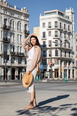 full length of brunette woman with straw hat looking away on urban street