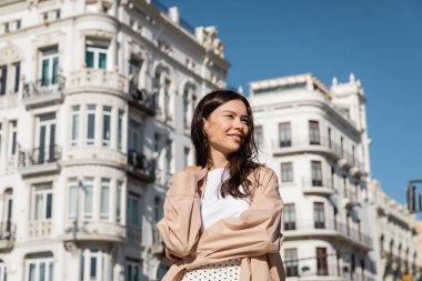 happy woman in beige shirt looking away while standing on urban street