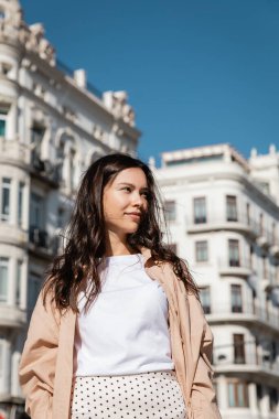 brunette woman smiling while standing on urban street and looking away 
